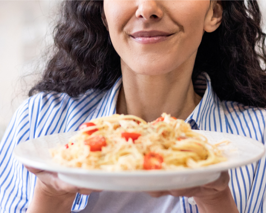 A woman holds a plate of pasta with cherry tomatoes, wearing a blue and white striped shirt, smiling slightly.