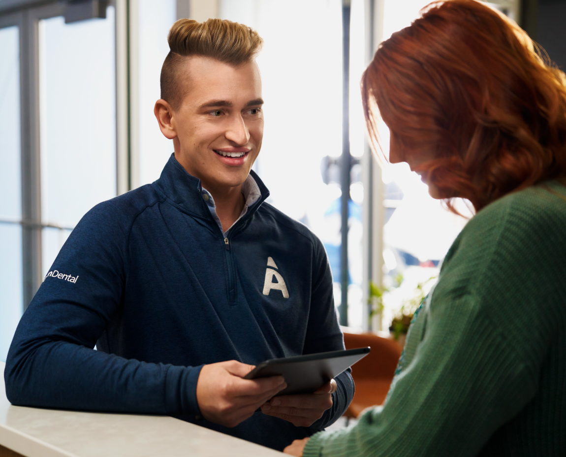 A young dental assistant in a blue jacket with Aspen Dental 'A' logo, holding a tablet and smiling at a woman with red hair who is looking at the tablet, in a brightly lit indoor setting.