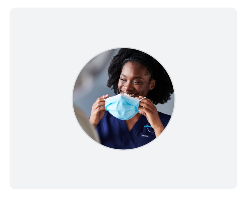 A woman in medical scrubs is smiling and holding a surgical mask.
