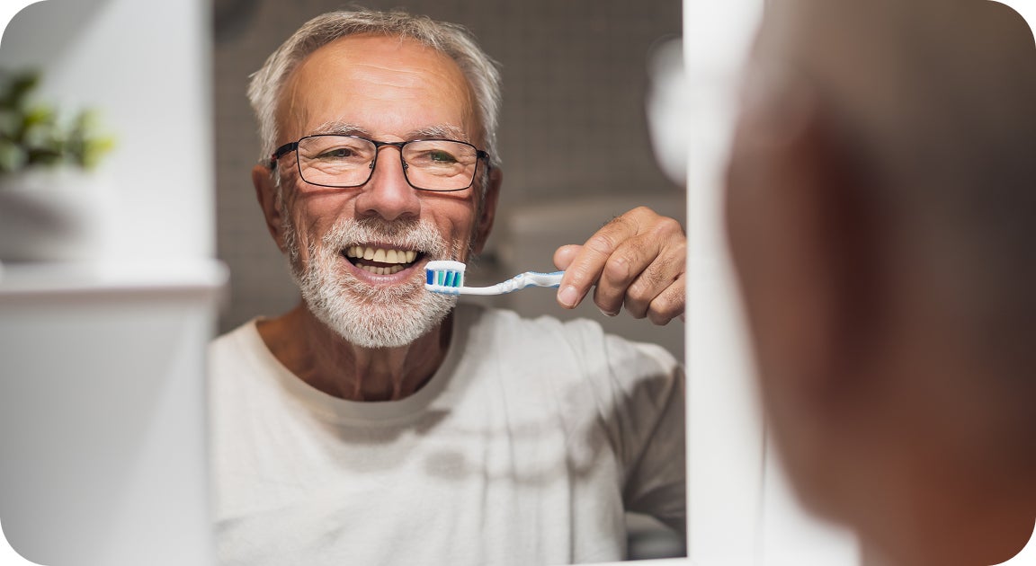 An older man smiles while brushing his teeth in the mirror, showing how maintaining dental hygiene supports the long-term success of implants in seniors. This visual reinforces that with proper care, dental implants can remain safe and effective at any age.