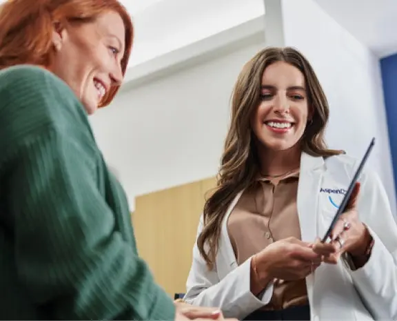 A friendly dental professional in a white coat discussing treatment options with a smiling female patient, using a tablet for patient education in a modern dental office setting.