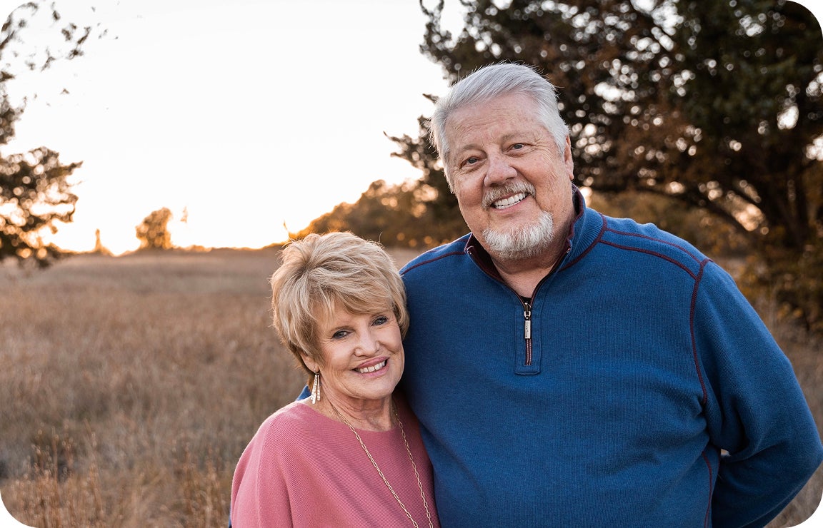 An older couple stands smiling together in a field at sunset, showing restored confidence after addressing delaying tooth replacement. Their natural expressions and relaxed posture reflect improved quality of life.