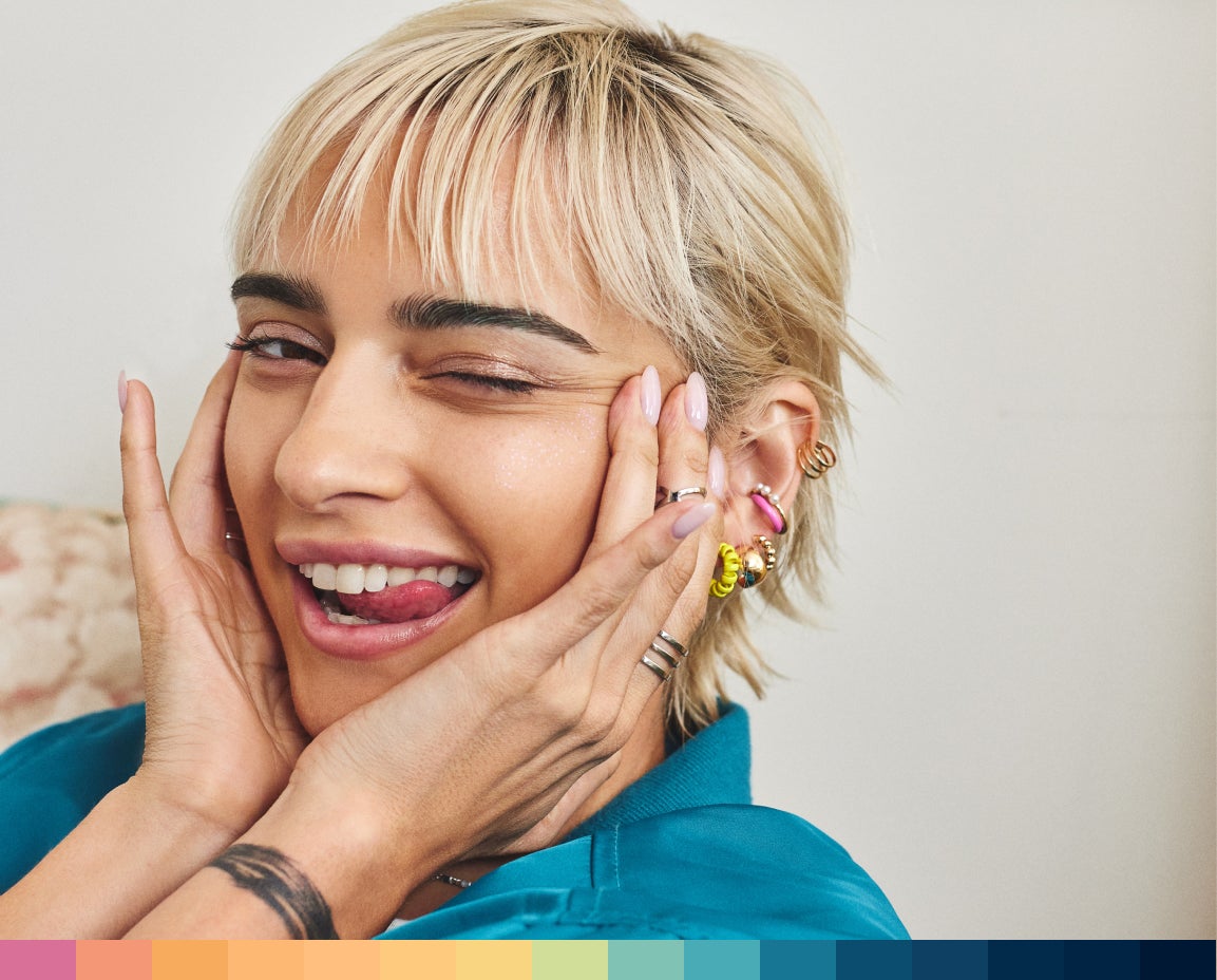  A woman winking with short blonde hair, hands on cheeks, smiling.