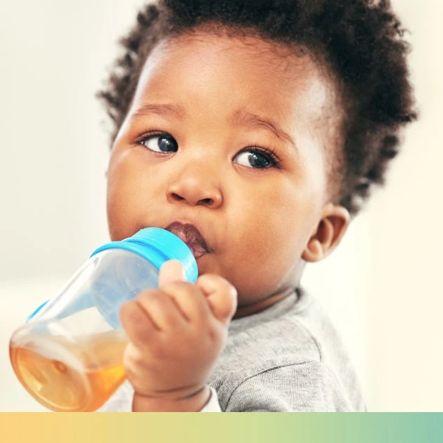 A toddler with curly hair drinks from a sippy cup with blue cover and clear cup, and filled with orange liquid.