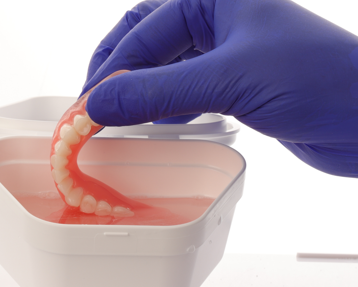 A gloved hand holding a set of dentures being immersed in a container filled with dentures cleaning solution.