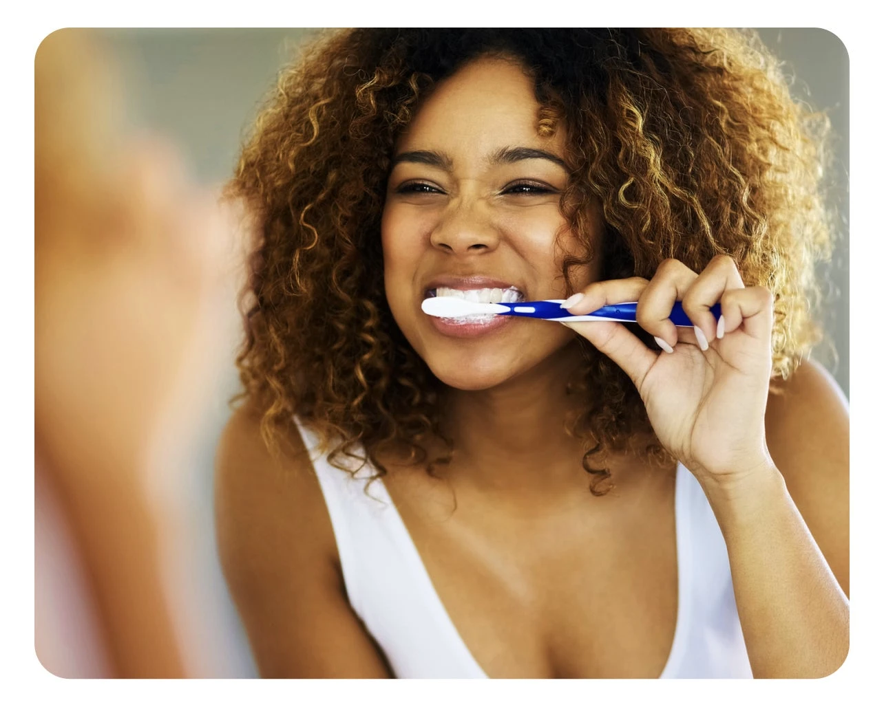 A woman with curly hair is brushing her teeth with a blue toothbrush, smiling and looking into a mirror.