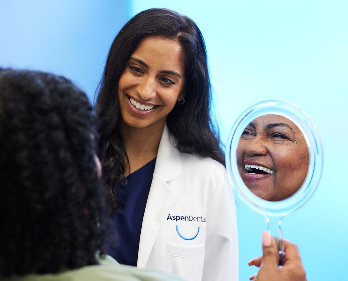 A smiling patient holds up a mirror reflecting her teeth while a dentist in an Aspen Dental coat looks on. Captures a positive post-treatment moment associated with dental fillings.