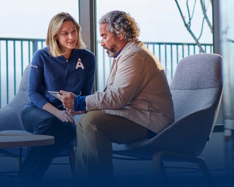 A man and a woman are seated and conversing; woman is wearing blue sweatshirt with an 'A' logo and is showing something on a tablet to the man. They are inside a modern setting with a railing and windows in the background.