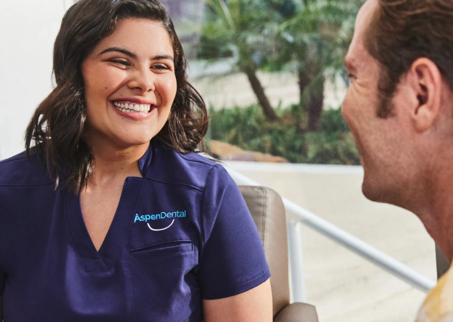 Smiling woman in a navy blue scrubs, with an Aspen Dental logo, speaks to a seated man in an indoor setting with plants in the background.