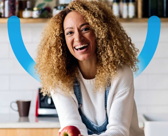 Woman with curly hair smiling and holding an apple in a kitchen, symbolizing dental confidence and health.