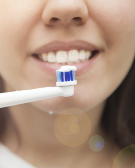 Close up of a smiling person with a toothbrush near their teeth, toothpaste on, with lens flare bubbles visible.