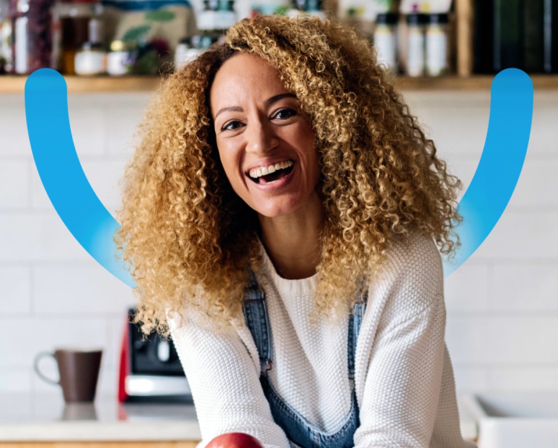 A picture of a woman with beautiful curly hair that's blonde, smiling brightly. Behind her is a kitchen counter with a coffee mug. An Aspen Dental blue smile graphic is shown layered behind her.