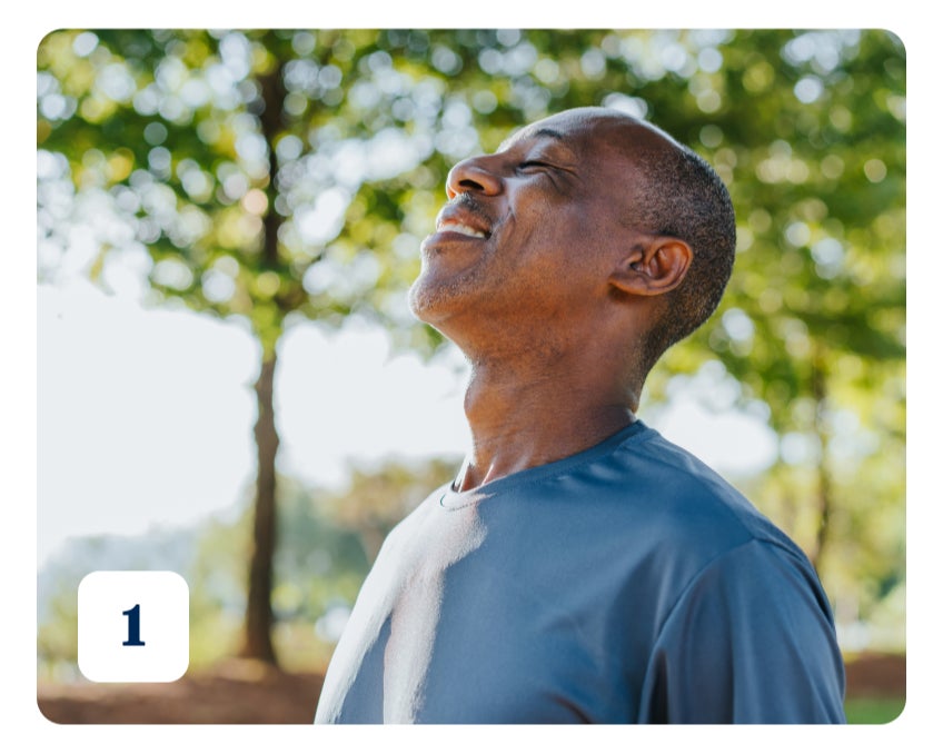 A man with his eyes closed and in a cobalt blue shirt, pointing his face up to the sky. In the background there are green trees and sunny skies.