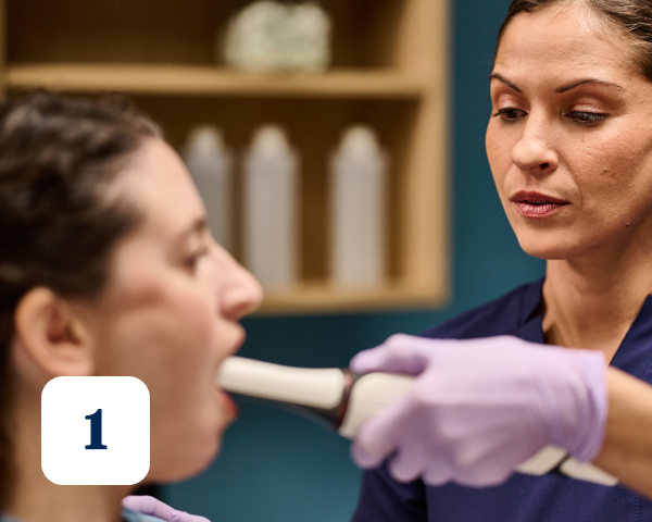 A dental professional in gloves examines a patient's open mouth with a medical instrument. Number "1" is displayed in the bottom-left corner.