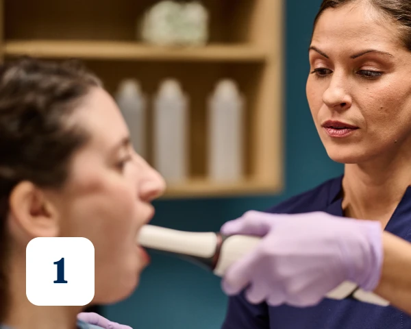A dental professional in gloves examines a patient's open mouth with a medical instrument. Number "1" is displayed in the bottom-left corner.