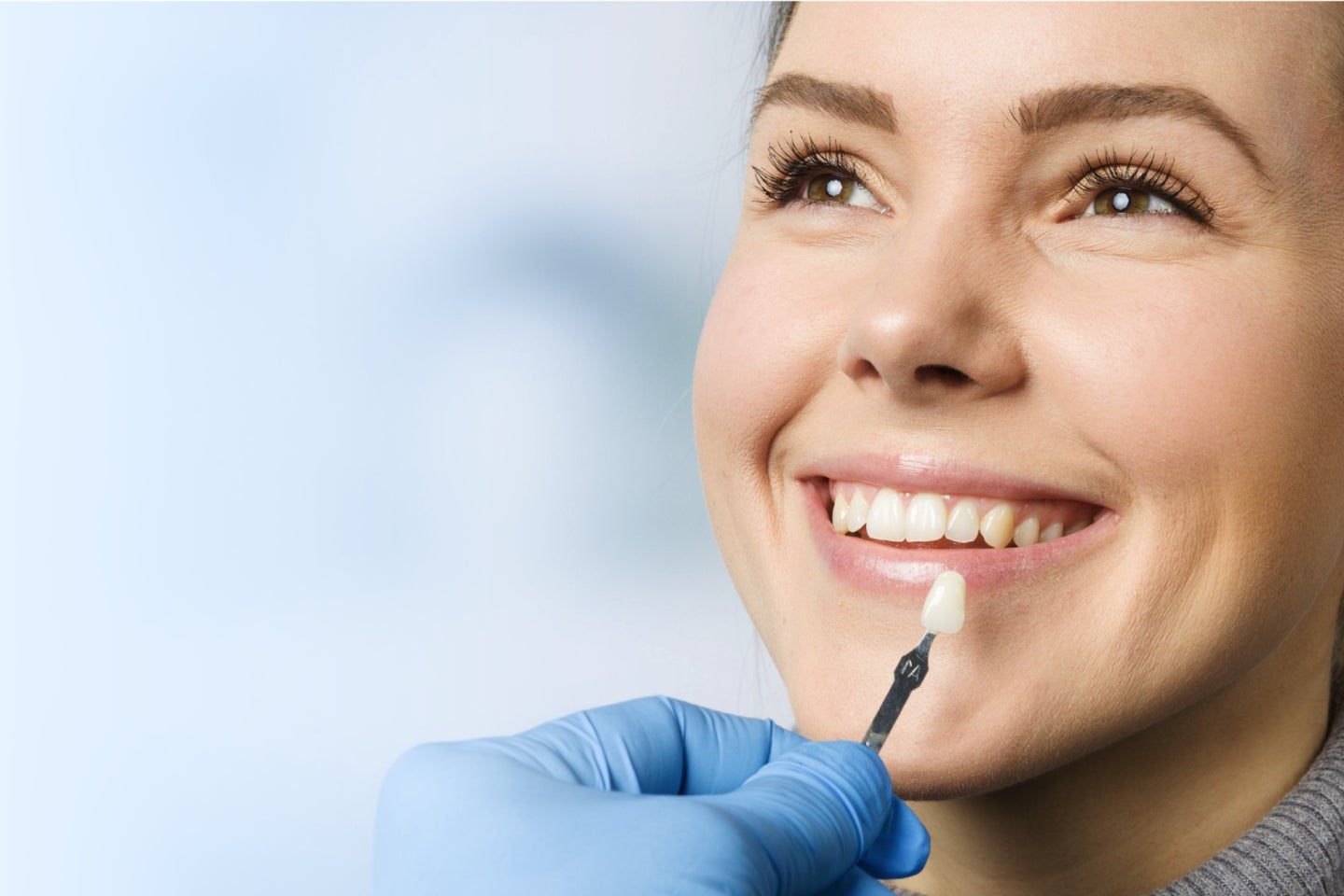 A dentist in a blue glove holds a single tooth shade guide near a smiling woman's teeth, matching the color for the veneers.