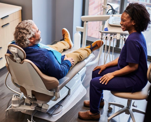 Aspen Dental patient seated in a dental chair having a friendly discussion with a dental assistant in a modern treatment room.