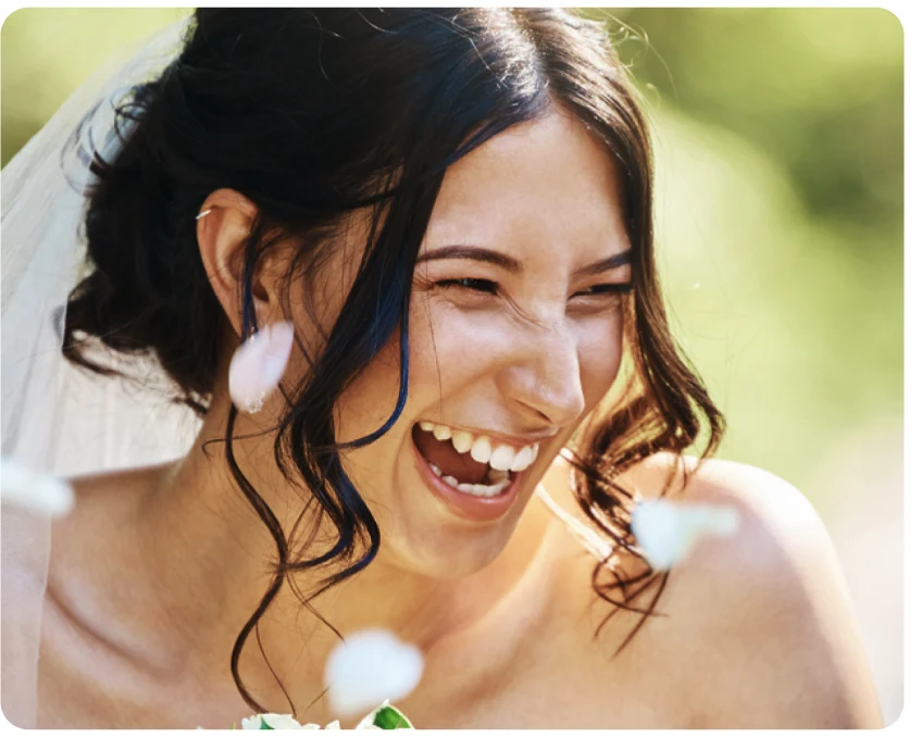 A bride wearing a veil, leaning forward and laughing. White flower petals are flying in the air.