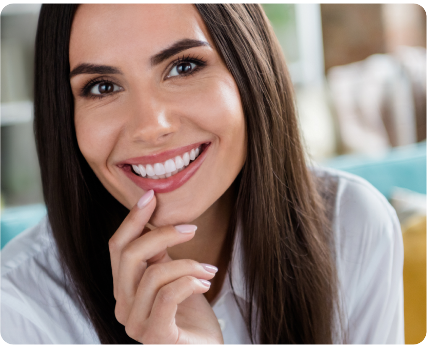 A woman with long dark hair smiles, resting her hand gently on her chin. She is wearing a white shirt and is seated indoors, exuding confidence possibly from impeccable veneers.