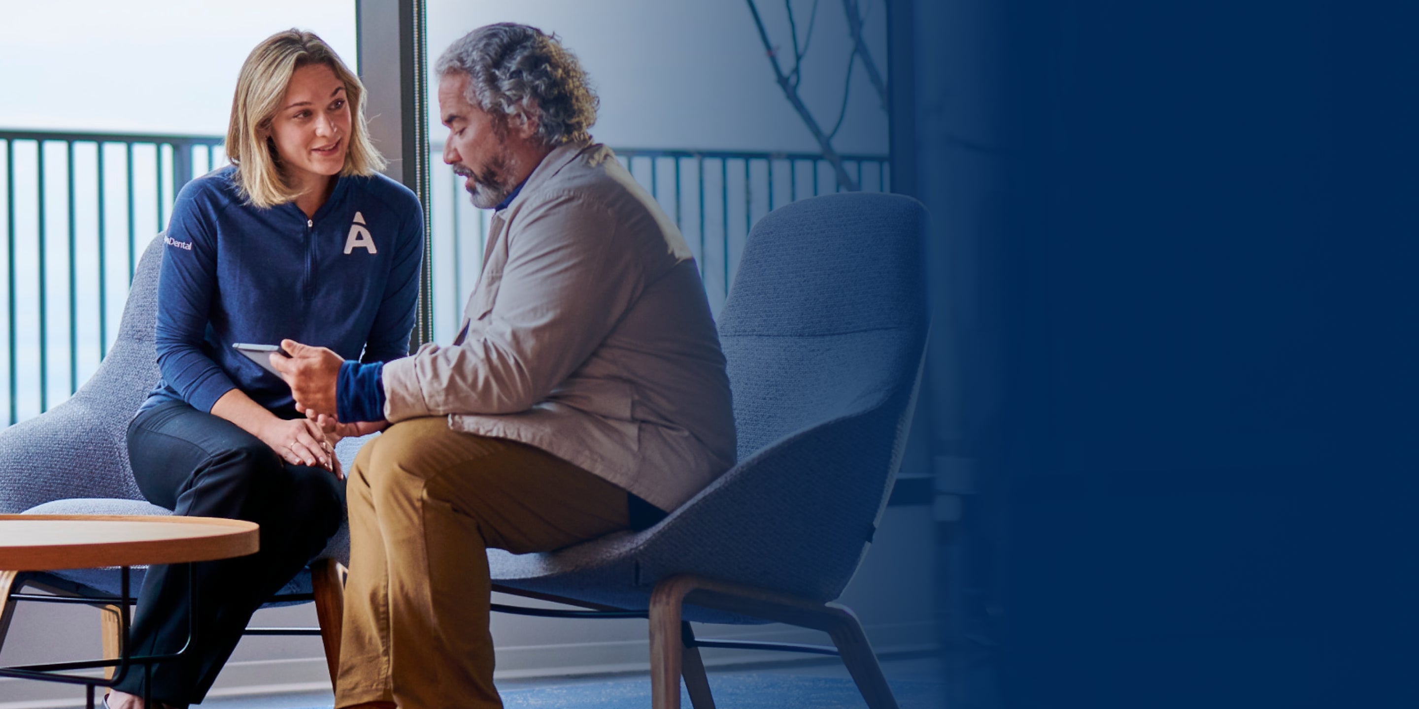 A man and a woman are seated and conversing; woman is wearing blue sweatshirt with an 'A' logo and is showing something on a tablet to the man. They are inside a modern setting with a railing and windows in the background.