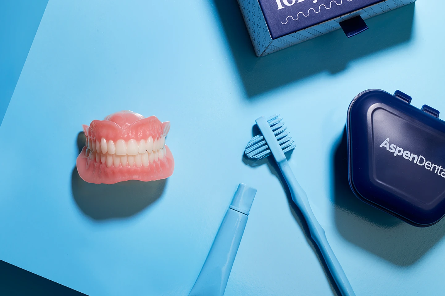 A denture model, denture storage case, toothbrush and toothpaste laid on a blue table representing a package included for new denture-wearer for Flexilytes℠ partial dentures.