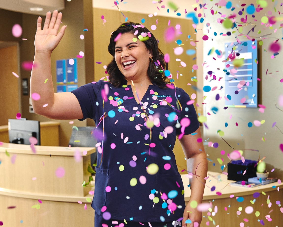 A woman in a blue Aspen Dental scrubs smiling and waving amid colorful confetti in a brightly lit room with office furniture in the background.