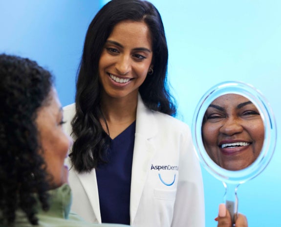 A patient at Aspen Dental smiles while holding a mirror, reflecting their happiness after a dental veneers procedure, with a caring dentist standing nearby.