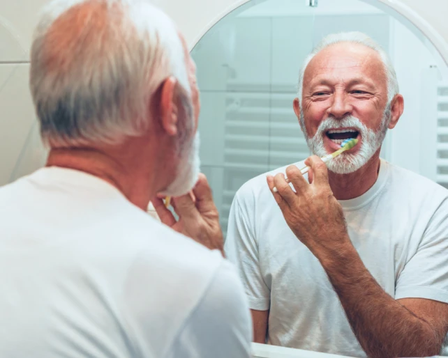 An older man brushing his teeth while looking in the bathroom mirror to prevent periodontal disease.