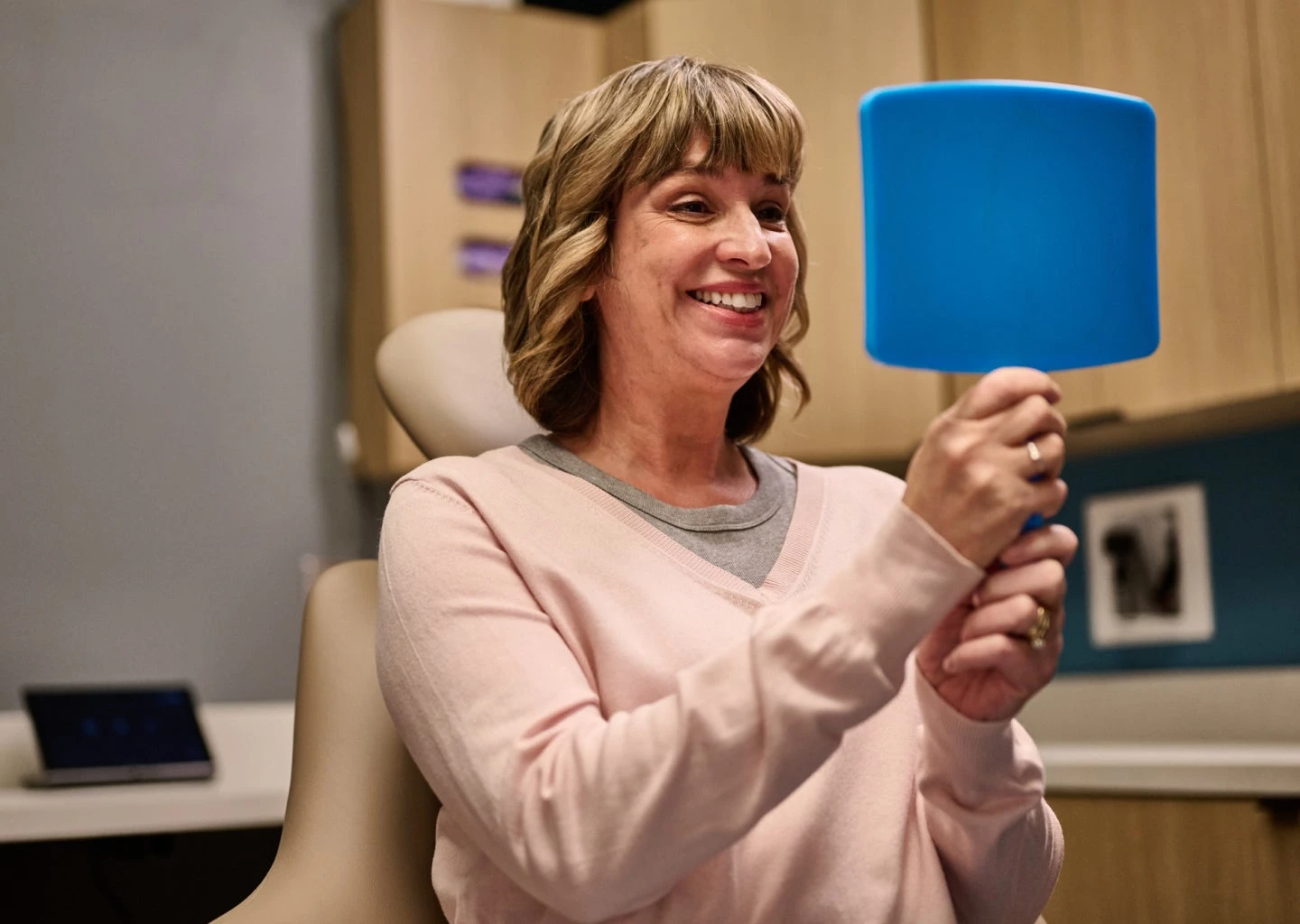 A woman with shoulder-length hair holding a blue hand mirror and smiling while sitting in an office setting with wooden cabinets and a laptop in the background, showcasing her new dental bridge.