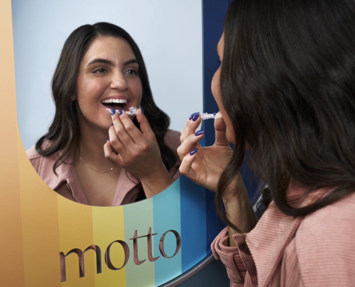 A woman patient standing in front of a mirror in the Motto office and placing clear aligners on her teeth.