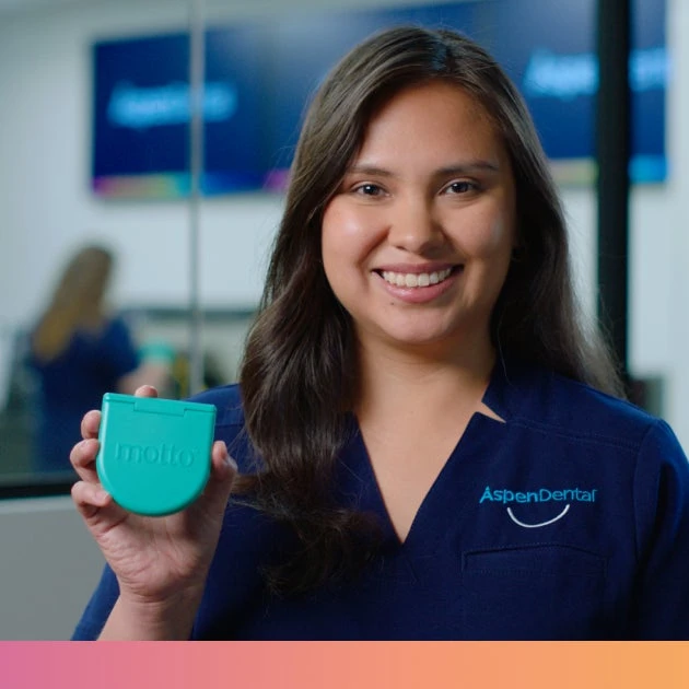 A woman in blue scrubs is smiling and holding a turquoise Motto clear aligners case.