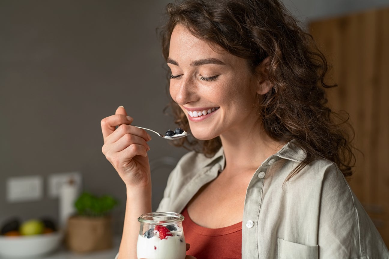 Smiling woman enjoying a jar of yogurt in a kitchen, symbolizing the confidence and comfort of a healthy smile after dental care from Aspen Dental.