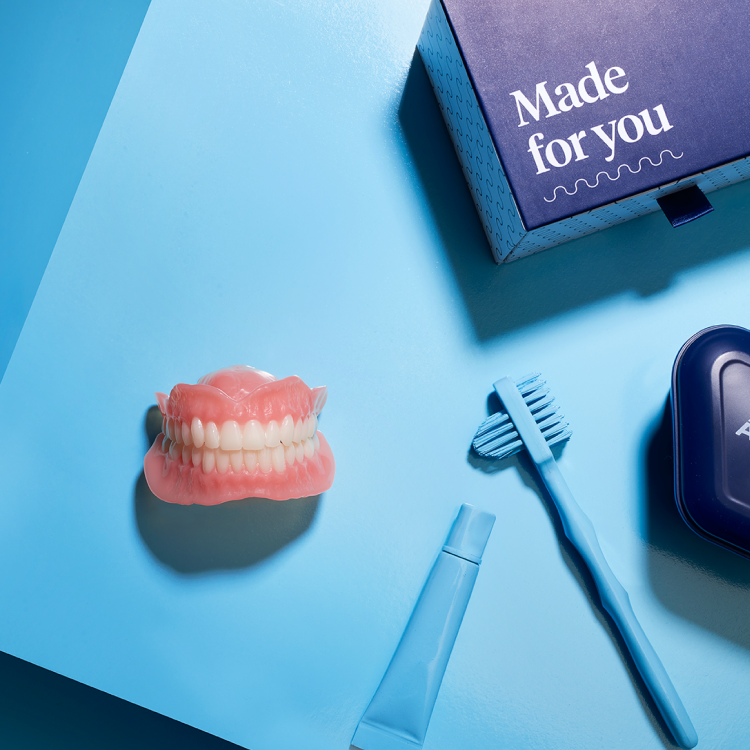 A denture model, denture storage case, toothbrush and toothpaste laid on a blue table representing a package included for new denture-wearer for Flexilytes℠ partial dentures.
