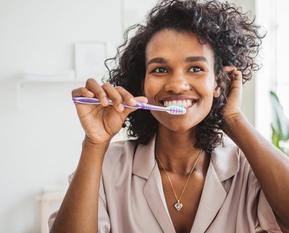 Smiling woman brushing her teeth in a bathroom, promoting good dental hygiene, with accompanying text discussing the importance of brushing twice a day and flossing to maintain enamel strength and prevent dental issues.