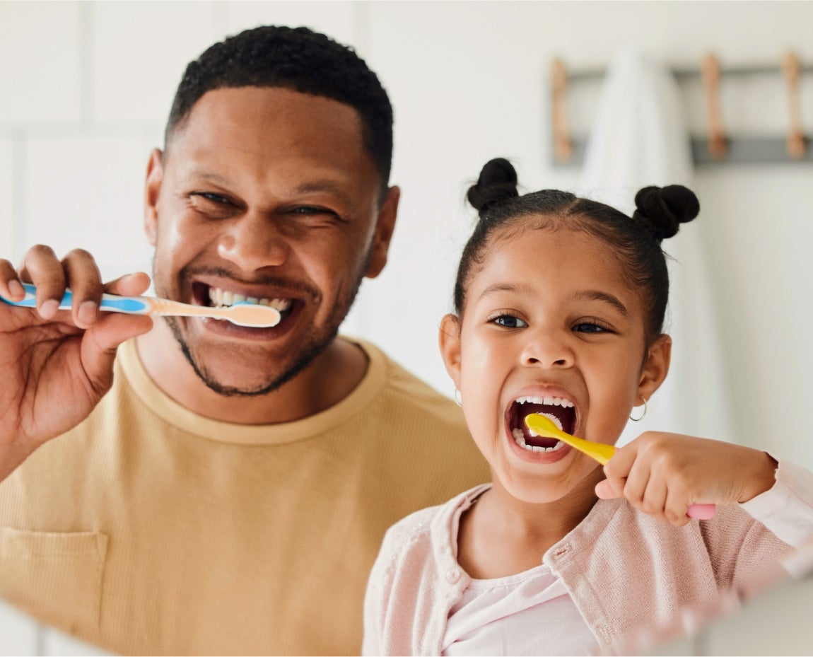 Smiling father and young daughter brushing their teeth together in a bathroom, promoting family dental hygiene and healthy habits.