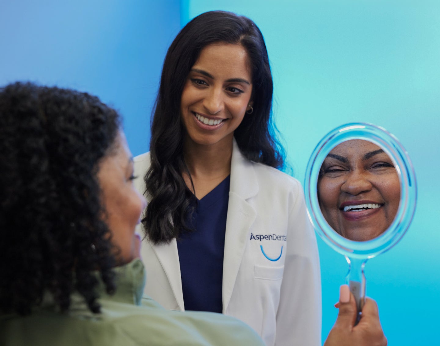 A happy Aspen Dental patient smiling into a handheld mirror, with a dentist in a white coat standing beside her, both reflecting satisfaction with the dental care on a bright blue background.