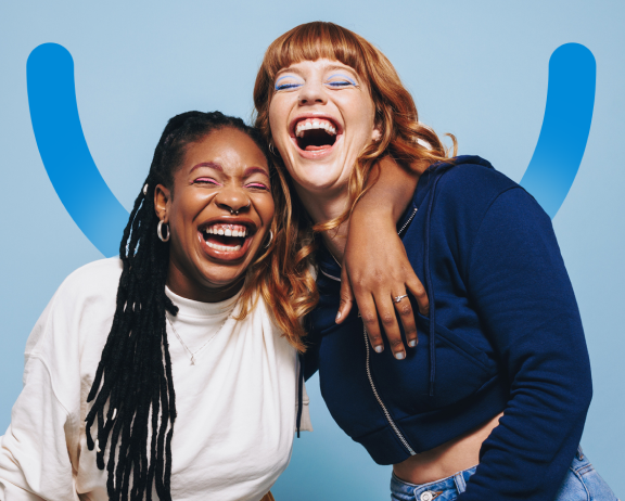Two women joyfully laughing in front of a blue background with Aspen Dental smile icon, sharing a moment of happiness.