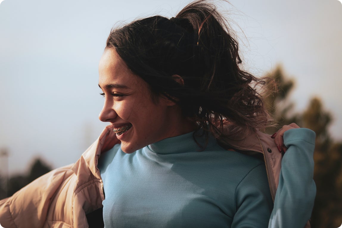 Young woman smiling outdoors while adjusting her jacket, showing braces and a confident, natural smile.
