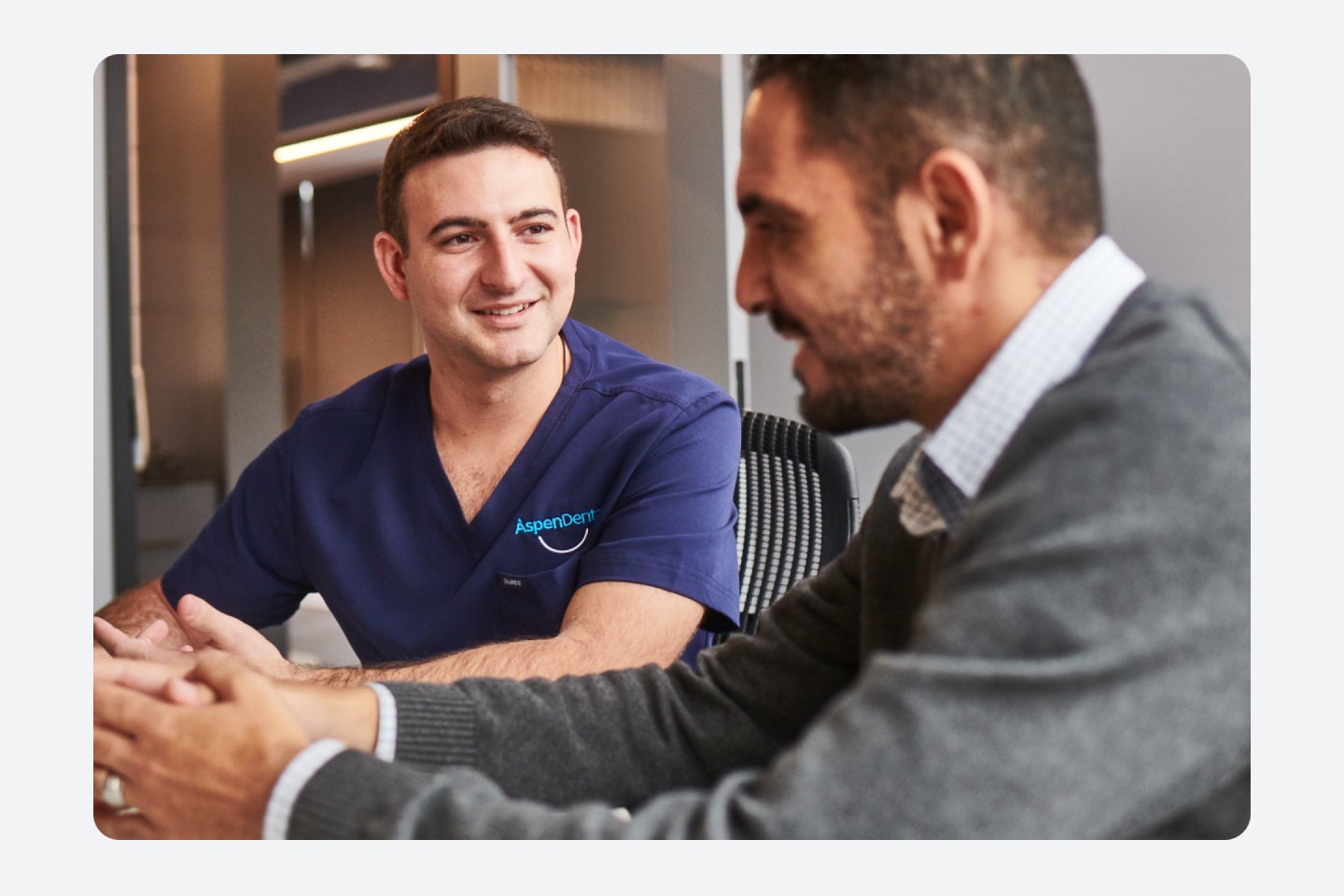An image of an Aspen Dental care team member wearing dark blue scrubs, consulting with a patient, who is smiling, with his hands folded together.