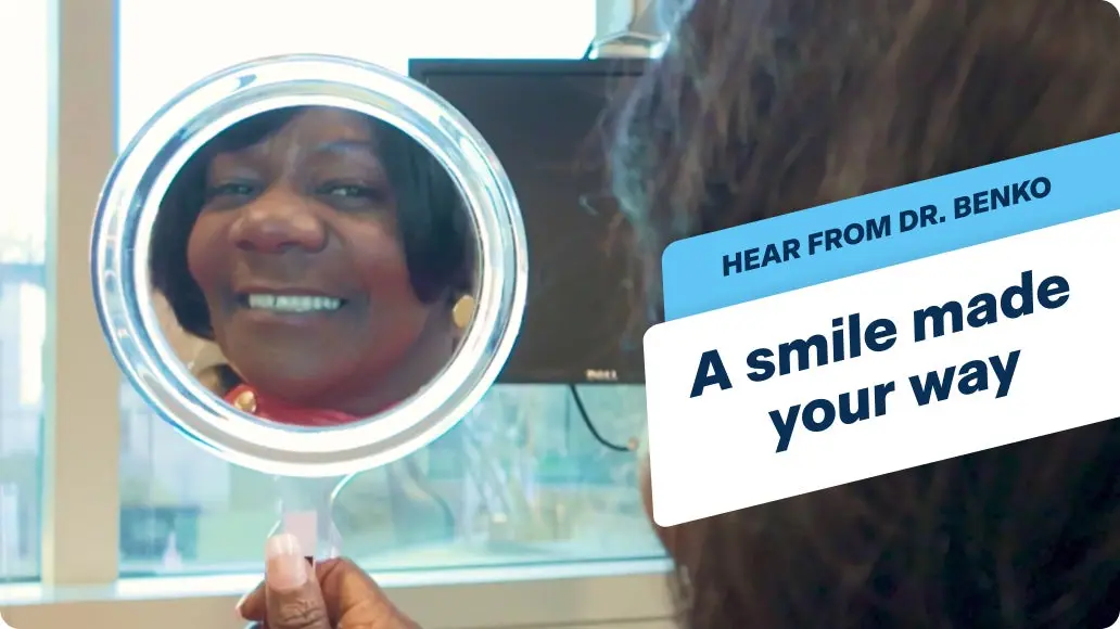 An image of a female patient admiring her new dental implants in a hand held mirror, with text: Hear from Dr.Benko, a smile made your way.