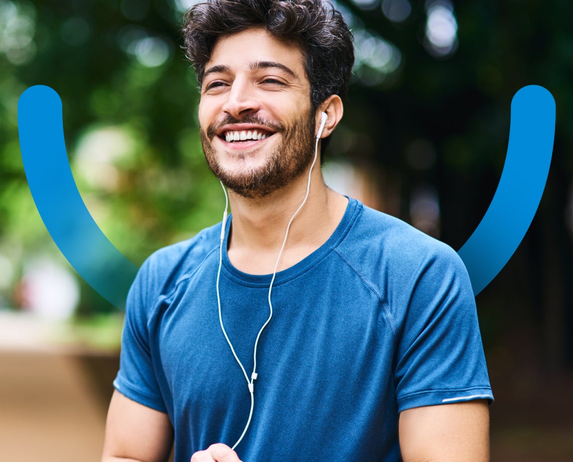 A man with curly hair and a beard, wearing earphones and a blue T-shirt, smiles while standing outside with blurred greenery in the background. Two curved blue lines are superimposed behind him to create a smile.