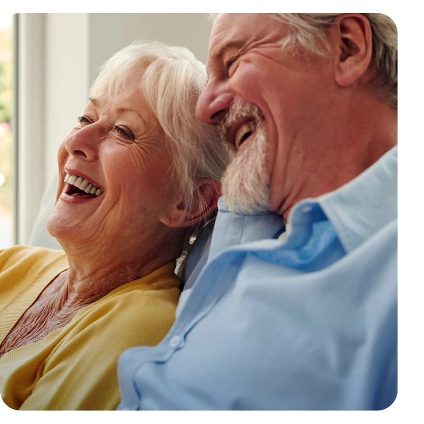 An elderly woman and man with gray hair are sitting closely together, smiling and laughing, possibly discussing pre-prosthetics tooth extractions for dentures and implants. Both are wearing casual clothes; the woman is in yellow, and the man is in blue.