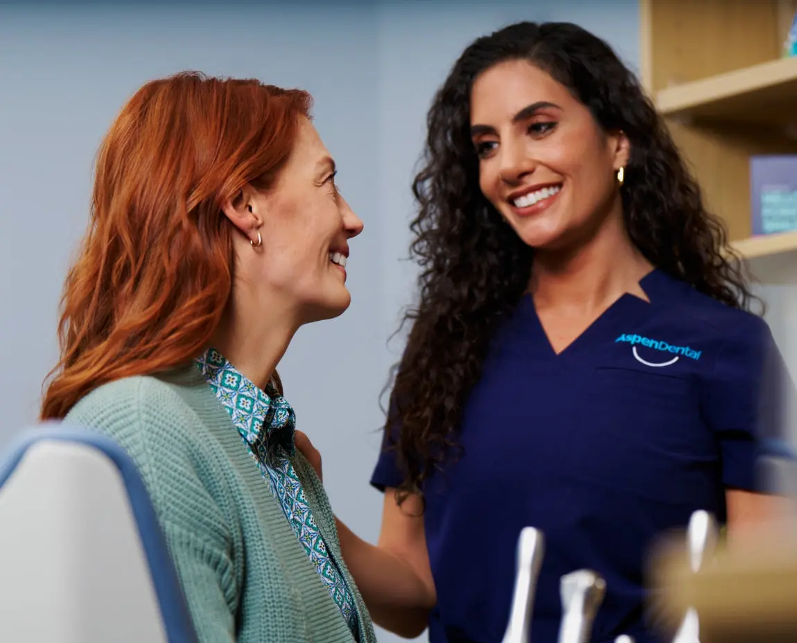 A dental professional in Aspen Dental scrubs smiles while speaking with a seated patient who has red hair and a green sweater.