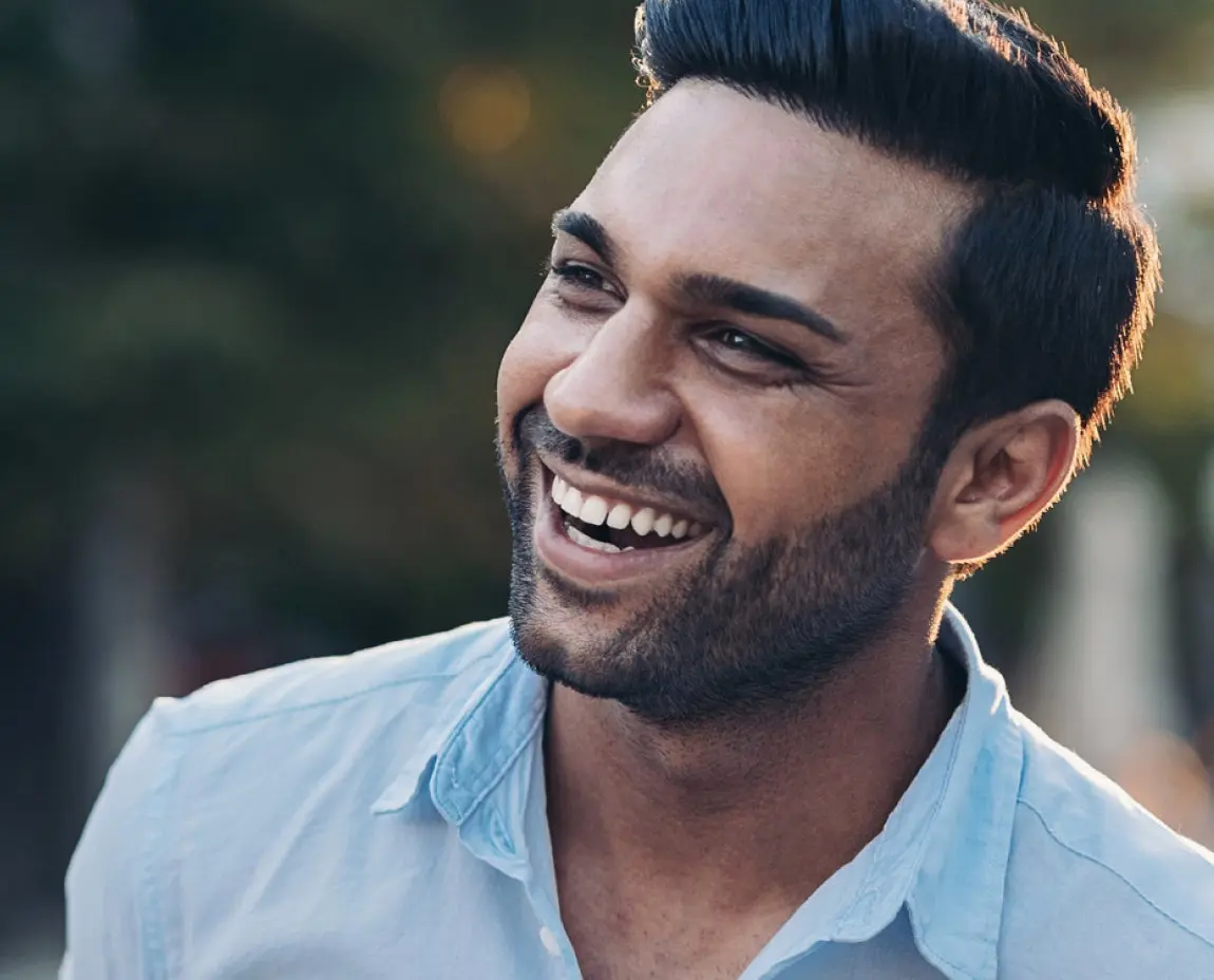 A man with short dark hair and a trimmed beard smiling while looking to his left. He is wearing a light blue button-up shirt and standing outdoors with a blurred green background.