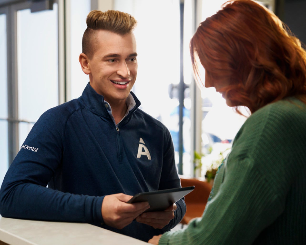 A man in a blue sweatshirt with 'A' logo and a woman with red hair converse over a tablet at a reception desk in a well-lit room.