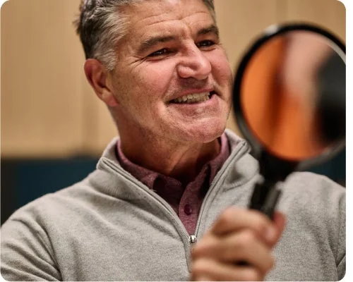 A smiling man with gray hair is looking at a magnifying mirror, wearing a gray sweater over a maroon shirt.
