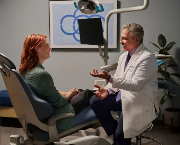 A friendly dentist in a white coat having a positive consultation with a female patient in a modern dental office. The patient is seated in the dental chair, engaged in conversation, indicating a welcoming and comfortable dental care experience.