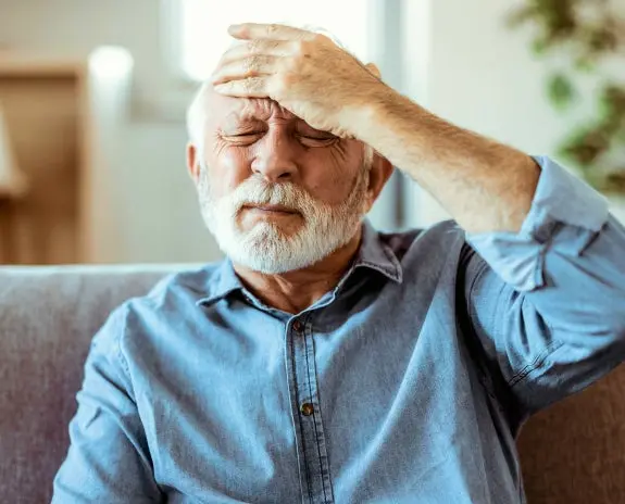 Elderly man sitting on a couch, holding his forehead in pain, illustrating the potential link between toothaches and headaches.