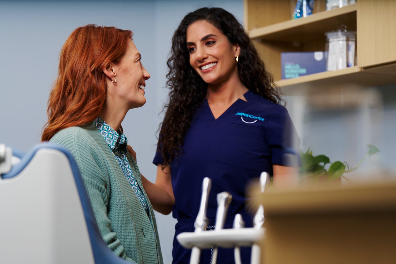 A smiling Aspen Dental team member in a purple uniform warmly engaging with a patient during a consultation in a dental office setting.