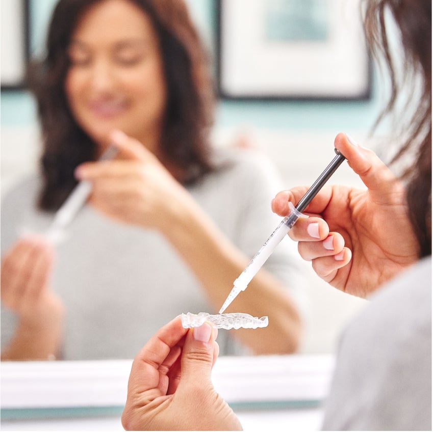 A woman uses her teeth whitening kit in a bathroom mirror. 